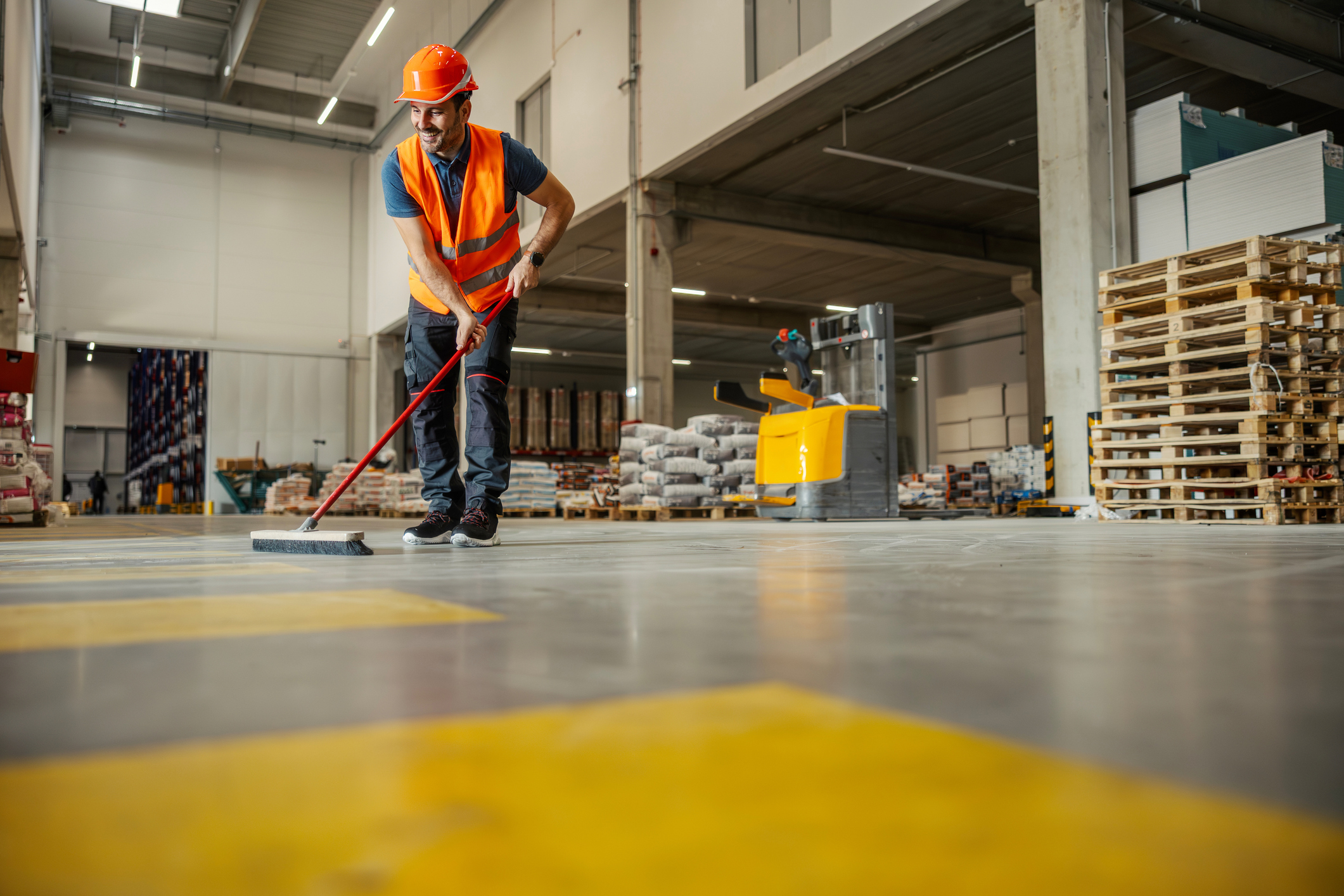 Smiling male worker in high-visibility vest and hardhat sweeping a warehouse floor, ensuring clean work environment and operational safety